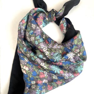 May include: A square scarf tied in a knot, featuring a vibrant floral pattern with shades of blue, purple, pink, and green on one side, and a solid black fabric on the reverse side. The floral print consists of small, dense blossoms.