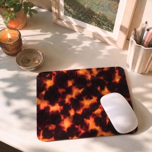 May include: A rectangular mousepad with a tortoiseshell pattern in shades of brown and orange. A white wireless mouse rests on the pad. The scene includes a lit candle, a small bowl with jewelry, and a framed picture.