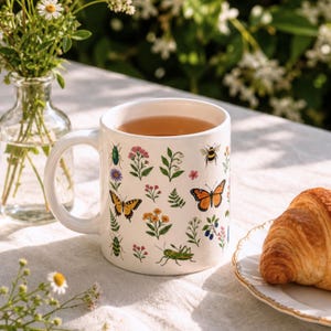 Peut inclure: Mug en céramique blanche orné d'illustrations colorées de papillons, d'abeilles, de fleurs et d'insectes. La tasse, remplie de thé, est posée sur une nappe blanche à côté d'un croissant sur une assiette. Un petit vase de fleurs est en arrière-plan.