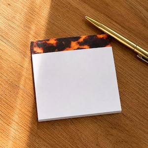 May include: A white notepad with a tortoiseshell pattern at the top edge. A gold pen rests on a wooden surface next to the notepad. The notepad is rectangular and appears to be unused.