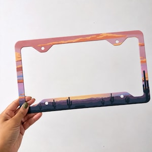 May include: A rectangular license plate frame featuring a desert landscape graphic. The design displays a sunset with orange and pink clouds above dark purple mountains and black silhouettes of saguaro cacti. It has four mounting holes.