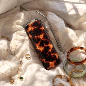 Puede incluir: Un vaso de acero inoxidable con un patrón de carey, una pajita plateada y joyas de oro, incluyendo collares, un anillo y pulseras. Los artículos están dispuestos sobre una superficie blanca texturizada.