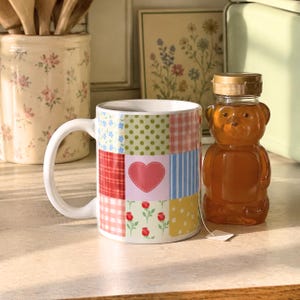 Op de afbeelding: Een mok met patchwork-ontwerp met een roze hart en bloemaccenten staat naast een honingbeerfles. De mok heeft verschillende patronen, waaronder stippen, strepen en ruitjes. Een theezakje is bevestigd aan de honingbeerfles.