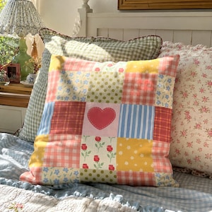 Peut inclure: Un coussin carré avec un motif patchwork composé de divers carrés de tissu en jaune, rose, bleu et vert. Les motifs incluent du vichy, des rayures, des pois et des fleurs, avec un carré central rose orné d'un cœur rouge.