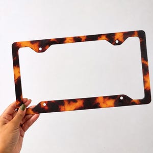 May include: A rectangular license plate frame featuring a mottled tortoiseshell pattern in shades of amber, orange, and dark brown. The frame has four circular mounting holes positioned at the corners to allow for secure attachment to a vehicle.
