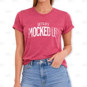 May include: A red t-shirt with rolled-up sleeves and the text "LET'S GET MOCKED UP" in white, distressed lettering. The model is wearing blue jeans and a gold necklace. The shirt is a solid color.