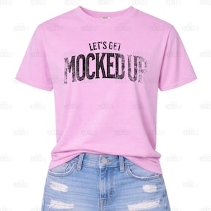 May include: A light pink t-shirt with the text "LET'S GET MOCKED UP" in distressed black lettering. The shirt has short sleeves and a crew neck. The shirt is paired with blue denim shorts.