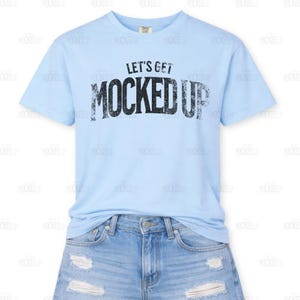 Puede incluir: Una camiseta azul claro con el texto "LET'S GET MOCKED UP" en letras negras desgastadas. La camiseta se combina con pantalones cortos de mezclilla. La camiseta es de cuello redondo.
