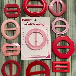 May include: A collection of various red and pink belt buckles in circular, octagonal, and rectangular shapes. One light pink circular buckle is attached to a white cardboard display card labeled Lansing Styles By A great name in Buttons Washable.