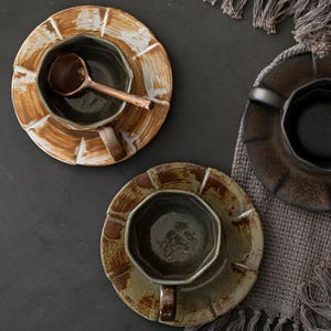 May include: Overhead view of three ceramic teacups and saucers. The cups are dark green with brown handles, and the saucers have a textured, earthy design in brown and white. One cup holds a small wooden spoon. A grey textile is in the background.