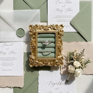 May include: A gold ornate frame holds two rings on a green velvet insert, surrounded by wedding stationery. The stationery includes invitations, envelopes, and a wax seal. A small bouquet of white roses and greenery is also present.