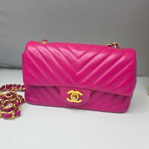 May include: A bright pink, quilted leather shoulder bag with a gold chain strap. The bag features a chevron pattern and a gold-tone clasp. The bag is a classic design, suitable for everyday use.