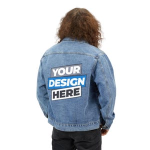 May include: A blue denim jacket featuring a rectangular graphic on the back with the text YOUR DESIGN HERE in white and blue lettering. The jacket has long sleeves, a collar, and visible stitching details across the back panel and shoulders.