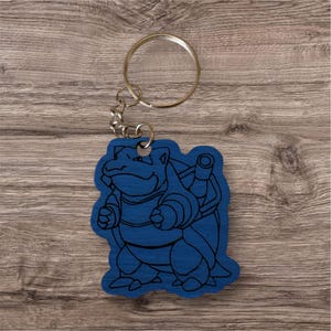 May include: A blue wooden keychain featuring a laser-engraved outline of the character Blastoise. The keychain includes a silver-toned metal split ring attached by a short chain, showcasing the creature's shell, cannons, and distinct body shape.
