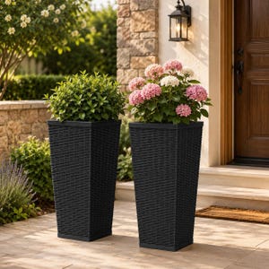 May include: Two tall, square, black planters with a woven texture. One planter contains a dense, green leafy shrub, while the other holds a flowering plant with clusters of pink and white hydrangea blossoms, both standing on a stone patio.