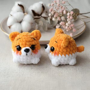 May include: Two handmade crochet dog toys. The toys are orange and white, with black eyes and pink cheeks. The toys are sitting on a white plate with cotton and flowers in the background.