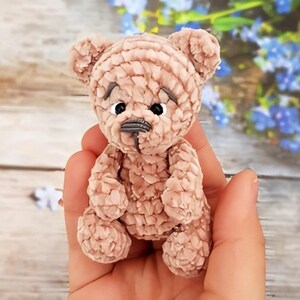 May include: A small, handmade teddy bear in a light brown color. The bear has a textured, plush appearance with visible stitching. It features small, black eyes and a stitched nose. The bear is held in a person's hand, with a blurred background of wooden planks and flowers.