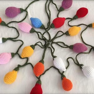 May include: A colorful crochet light string featuring various bulb colors, including orange, purple, pink, yellow, white, red, and blue. The bulbs are connected by green crocheted cords. The handmade string lights are arranged on a white surface.