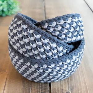 May include: Three handmade crochet baskets in a nested arrangement. The baskets are a combination of grey and white, with a textured pattern. The rims are a solid grey, and the baskets are sitting on a wooden surface.