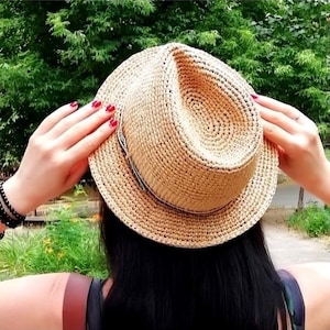 Op de afbeelding: Een geweven strooien fedora hoed met een donkere band. De hoed heeft een lichtbruine kleur en heeft een klassieke fedora vorm. De hoed wordt vastgehouden door een persoon met lang zwart haar en rode nagellak.