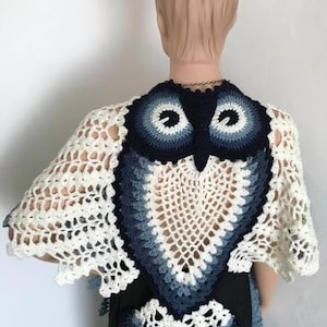 May include: A handmade crochet shawl shaped like an owl. The shawl uses white, blue, and navy yarns. The owl's face is at the back, with wings over the shoulders. The open weave design gives a delicate appearance. A unique, handcrafted item.