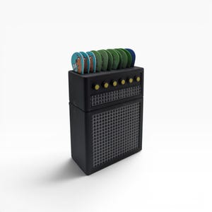 May include: A black guitar amplifier-shaped holder containing several guitar picks. The top section features six yellow control knobs and a grid pattern, while the bottom section has a larger speaker grille. Some picks display a cartoon dog graphic.