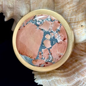May include: A round, gold-toned metal pendant with a large, polished stone center. The stone features a mix of pink, blue, and white patterns. The pendant is set against a seashell background, creating a natural and elegant aesthetic.