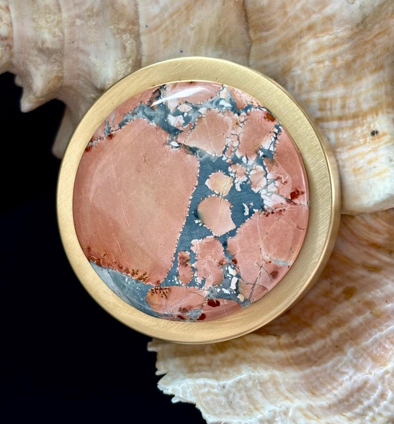 May include: A round, gold-toned metal pendant with a large, polished stone center. The stone features a mix of pink, blue, and white patterns. The pendant is set against a seashell background, creating a natural and elegant aesthetic.