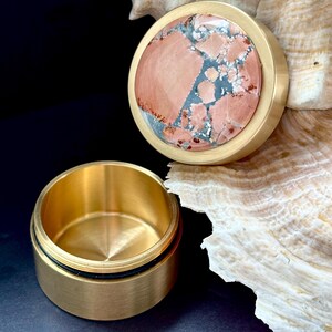 May include: A small, round, brass container with a lid. The lid features a circular stone inlay with a mix of pink, gray, and blue hues. The container is open, revealing its interior. The background is dark, with a seashell.
