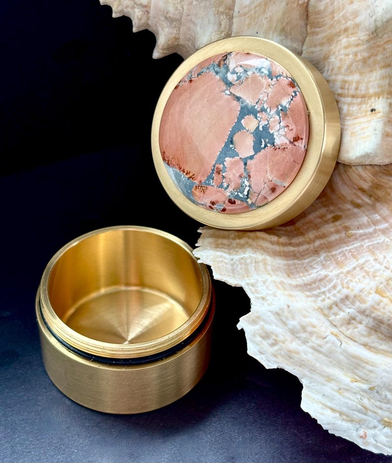 May include: A small, round, brass container with a lid. The lid features a circular stone inlay with a mix of pink, gray, and blue hues. The container is open, revealing its interior. The background is dark, with a seashell.