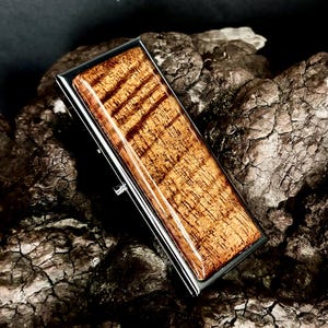 May include: A rectangular money clip featuring a polished dark metal frame and a smooth, glossy inlay of brown wood with a distinct, dark horizontal grain pattern. The object has a sleek, metallic edge and a reflective, finished surface.