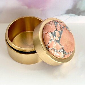 May include: A round, open brass box with a lid. The lid features a circular stone with a pink and gray pattern. The box is sitting on a white surface.