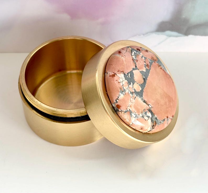May include: A round, open brass box with a lid. The lid features a circular stone with a pink and gray pattern. The box is sitting on a white surface.