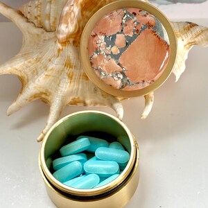 May include: A small, open, gold-colored pill container filled with light blue pills. The container has a matching lid with a stone top featuring a mix of pink, gray, and white patterns. A seashell is in the background.