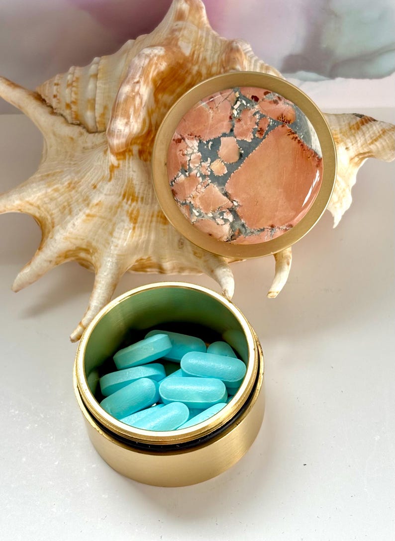 May include: A small, open, gold-colored pill container filled with light blue pills. The container has a matching lid with a stone top featuring a mix of pink, gray, and white patterns. A seashell is in the background.