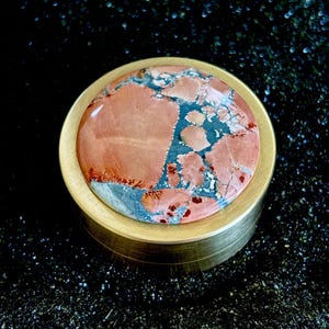May include: A round, brass-colored container with a lid featuring a polished stone. The stone has a mix of red, blue, and gray hues. The container is set against a dark, textured background.