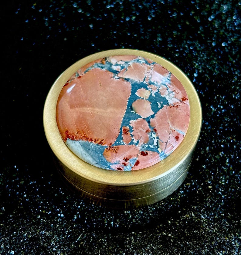 May include: A round, brass-colored container with a lid featuring a polished stone. The stone has a mix of red, blue, and gray hues. The container is set against a dark, textured background.