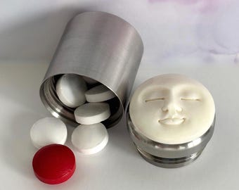 May include: A stainless steel pill container with a smiling face lid, next to a spilled assortment of white and red pills. The container is cylindrical and has a brushed metal finish. The pills are round.