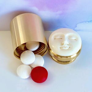 以下が含まれることがあります： 閉じた目と穏やかな表情の白い顔が彫刻されたスクリューキャップ付きの、円筒形の金色の金属製ピルケース。開いた容器からは、いくつかの白い丸い錠剤と1つの赤い丸い錠剤がこぼれ落ちています。