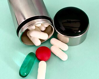 May include: A stainless steel pill container with a black lid, open to reveal white capsules. Several pills, including a red tablet and a green capsule, are scattered around the container on a light blue surface. The container is cylindrical.