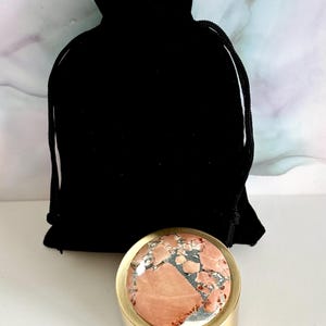 May include: A small, round, gold-colored container with a patterned, pink and gray stone top. A black velvet drawstring bag is behind the container. The stone has a mix of colors and textures, adding a unique touch to the item.