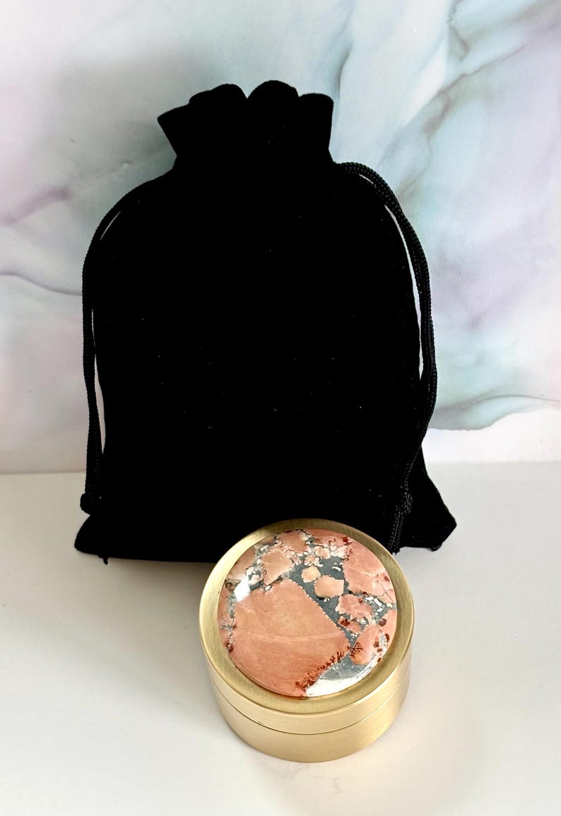 May include: A small, round, gold-colored container with a patterned, pink and gray stone top. A black velvet drawstring bag is behind the container. The stone has a mix of colors and textures, adding a unique touch to the item.