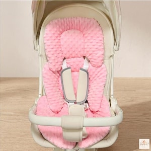 May include: A pink, plush baby stroller insert with a textured surface. The insert has a headrest and a seat with a harness. The stroller frame is off-white with a curved handle.