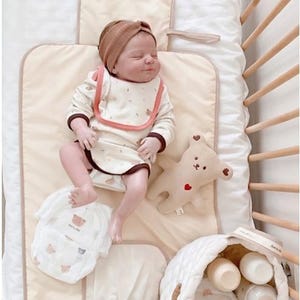 May include: A sleeping baby wearing a brown headband and a cream-colored outfit with a bear design, lying on a beige changing pad. A beige teddy bear with a red heart, diapers, and a quilted basket with bottles are also visible.