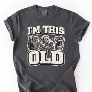May include: Dark grey t-shirt with the text "I'M THIS OLD" in white block letters. The graphic includes illustrations of car parts, such as a distributor cap, a points set, and a choke. The word "CHOKE" is also visible.