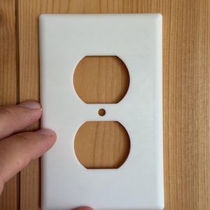 May include: A rectangular white plastic electrical outlet wall plate featuring two stacked hexagonal openings for a duplex receptacle and a small centered circular hole for a mounting screw, designed to cover standard wall electrical outlets.