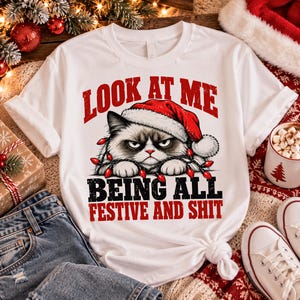 Peut inclure: Un t-shirt blanc à manches courtes avec un motif de chat grincheux portant un bonnet de Père Noël rouge et emmêlé dans des guirlandes lumineuses rouges. Le texte indique LOOK AT ME BEING ALL FESTIVE AND SHIT en lettres rouges et noires.