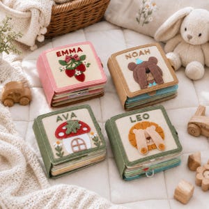 May include: Four square fabric activity books with felt appliques and embroidered names. One features strawberries and 'EMMA', one a bear and 'NOAH', one a mushroom house and 'AVA', and one a lion and 'LEO'. Each book has colorful pages.