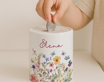 Peut inclure: Tirelire cylindrique en céramique blanche ornée d'un motif floral avec des fleurs et des papillons colorés. Le nom "Elena" est écrit en cursive. Une main insère une pièce dans la fente supérieure.