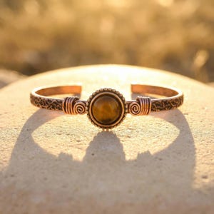 Könnte beinhalten: Kupferarmreif mit einem zentralen, runden, braunen Edelstein. Das Armband hat ein detailliertes, offenes Design mit Spiralakzenten auf beiden Seiten des Edelsteins. Das Metall hat einen warmen, rötlich-braunen Farbton.